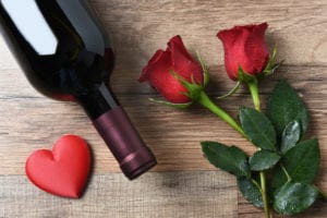 A wine bottle and two red roses and heart on a rustic wood table. Overhead view in horizontal format. Valentines Day Concept.